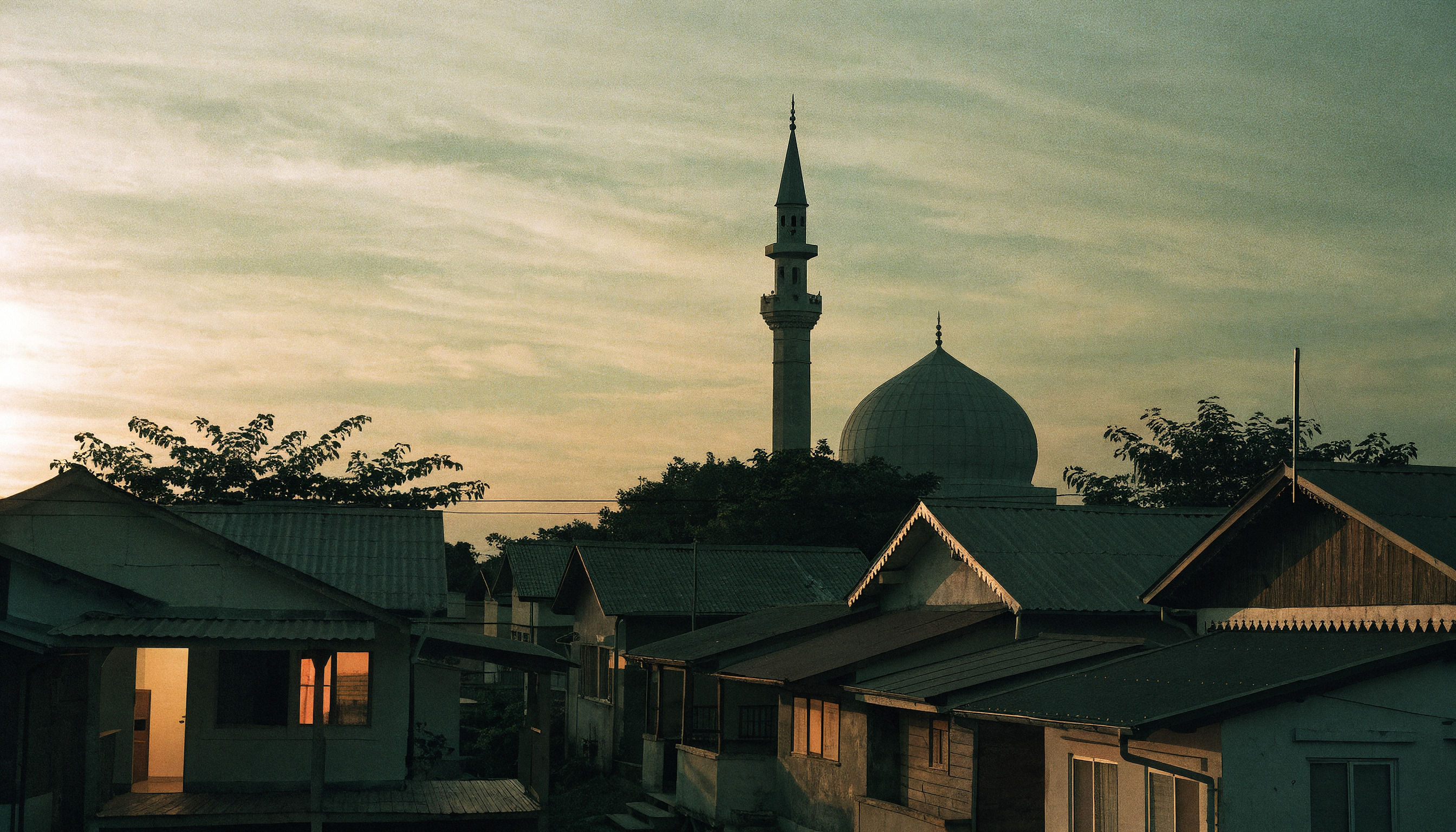 Alacakaranlıkta bir mahalle silüeti, cami minaresi ve sıcacık yanan ev pencereleri