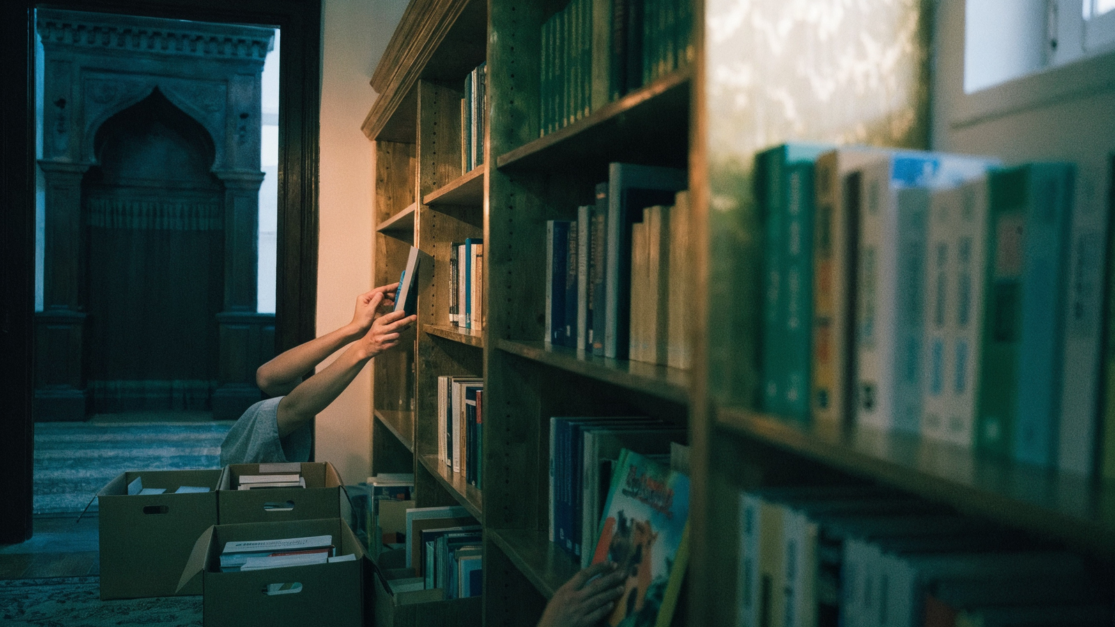 Cami yanındaki odada ahşap raflara kitap yerleştiren eller; kemerli kapı, koliler ve alt rafta kitaba uzanan çocuk eli. Yüz yok.